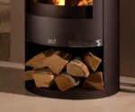 Alternativ tekst: Nærbillede af en moderne brændeovn med sort stålkabinet. Fokus er på brænderummet i bunden af ovnen, hvor træet er pænt stablet. Det varme lys fra ilden i brændeovnen giver billedet en hyggelig atmosfære. Perfekt til elskere af brændeovne, som sætter pris på et stilfuldt design.