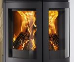 Billedet viser et detaljeret udsnit af en brændeovn med fokus på glasruden. Gennem den klare rude ses flammernes livlige spil tydeligt, hvilket kan skabe en hyggelig og varm atmosfære i rummet. Den elegante ramme om ruden understreger brændeovnens moderne design, der er ideelt til ethvert stilfuldt hjem.