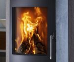 Das Bild zeigt einen Ausschnitt eines Kaminofens mit einem Fokus auf der Tür mit Sichtscheibe. Durch die große Glasscheibe ist ein lebendiges Flammenspiel zu sehen, das ein warmes und einladendes Ambiente vermittelt. Der Rahmen der Tür ist in einem eleganten, dunklen Farbton gehalten, der zur modernen Ästhetik des Kaminofens beiträgt. Der Ofen scheint robust verarbeitet zu sein und eignet sich perfekt für gemütliche Abende mit knisterndem Feuer.