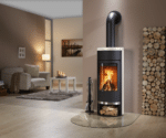 Een moderne houtkachel in een gezellige woonkamer met een brandend haardvuur. Ernaast een stapel hout, een bank en een salontafel met kaarsen.
