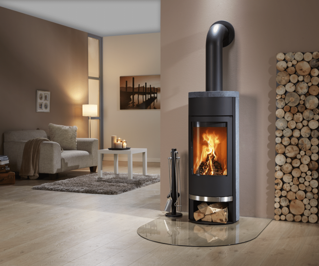 Filou Speckstein Kaminofen mit brennendem Feuer in modernem Wohnzimmer. Holzscheite im Fach darunter. Gemütliche Sitzecke mit Sofa, Teppich und Kerzen.