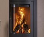 De focus van de foto ligt op een detail van de houtkachel, met name op de grote, heldere glasplaat. Deze biedt een indrukwekkend uitzicht op het laaiende vuur binnenin. De vlammen dansen levendig over de houtblokken en creëren een gezellige en uitnodigende sfeer. De gebogen vorm van de ruit zorgt voor een optimaal zicht op het vuur, waardoor deze houtkachel niet alleen functioneel is, maar ook esthetisch aantrekkelijk. Deze kachel combineert een modern design met een efficiënte warmteverdeling.