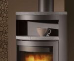Billedet viser den øverste detalje af et moderne komfur, nærmere bestemt området over glasdøren, hvor en grå kop er placeret på en hylde. Brændeovnen er lavet af stål og støbejern, hvilket giver den et elegant og moderne design. Et beroligende flammespil skinner gennem glasdøren. Denne brændeovn er den perfekte stilfulde tilføjelse til enhver stue og skaber en hyggelig atmosfære. Ideel til alle, der er på udkig efter en brændeovn af høj kvalitet til deres hjem.