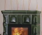 Das Bild zeigt einen Ausschnitt eines Kaminofens, genauer gesagt die detailreiche Gestaltung der Kachelverkleidung. Die grün gefliesten Kacheln verleihen dem Kaminofen ein elegantes und hochwertiges Erscheinungsbild. Im Fokus steht die präzise Verarbeitung und das klassische Design, das Wärme und Gemütlichkeit ausstrahlt. Durch die Glasfront ist das lebendige, wärmende Feuer sichtbar, was den Holzofen zu einem zentralen Blickfang macht. Dieser Kaminofen ist ideal für alle, die sowohl funktionale als auch ästhetische Merkmale schätzen.