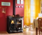 Op de foto is een elegante houtkachel te zien die in een gezellige woonkamer staat. De kachel heeft een robuust zwart oppervlak en oogt tegelijkertijd modern en klassiek. Hij bestaat uit twee delen, met een groot kijkvenster aan de linkerkant waardoor het vlammenspel zichtbaar is, en een tweede compartiment aan de rechterkant. De ruimte is warm en uitnodigend ingericht, met rode muren en gele gordijnen die het natuurlijke licht binnenlaten. Een mand met houtblokken staat naast de kachel en onderstreept de uitnodigende sfeer. De kachel is zowel een functioneel verwarmingselement als een stijlvol designelement dat de ruimte opwaardeert.