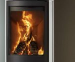 De afbeelding toont een detail van een houtkachel met een duidelijke focus op de gedetailleerd vormgegeven ruit. De vlammen in de kachel branden levendig en creëren een warme, uitnodigende sfeer. Het zwarte frame van de ruit geeft de houtkachel een moderne en elegante uitstraling. Ideaal voor stijlvolle woonruimtes waar gezellige uurtjes bij de houtkachel worden doorgebracht.