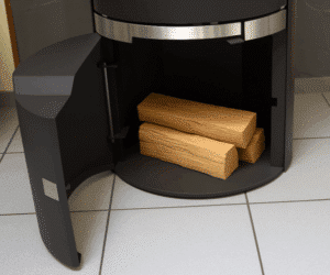 Das Bild zeigt das geöffnete Holzlager eines modernen Kaminofens, Modell Filou II HL. Im Inneren des Holzofens befinden sich mehrere Holzscheite. Die Konstruktion ist hochwertig, mit klaren Linien und einem eleganten schwarzen Finish. Besonders betont wird hier das praktische Holzfach, das für eine einfache Lagerung von Brennholz direkt am Kaminofen sorgt. Ideal für eine effiziente Heizlösung zu Hause.