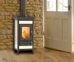 Beigefarvet brændeovn foran en stenmur, med synlige flammer og en glasdør, i et hyggeligt rum.