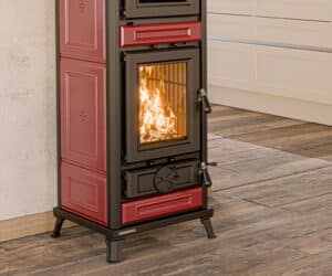 Rød Agnese Maiolica-ovn med brændende ild bag glas, står på trægulv foran hvid væg.