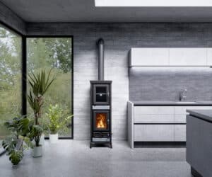 Moderne brændeovn i et lyst betonkøkken ved siden af store planter og høje vinduer, der lukker masser af lys ind.