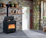 Wood-burning stove in a rustic room with an open flame, flanked by shelves and stone walls. Bright, inviting atmosphere.