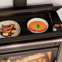 Wood-burning stove with open top plate; two plates with food are placed on it and a thermometer shows 300°C.