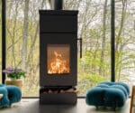 Modern wood-burning stove in dark design, shows flames through a glass front; behind it a large window front with autumn trees.