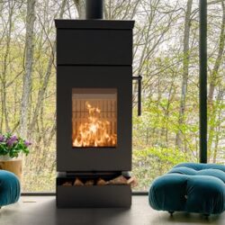 Modern wood-burning stove in dark design, shows flames through a glass front; behind it a large window front with autumn trees.