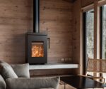 Modern wood-burning stove built into a wooden wall, flames visible. Cozy reading corner with sofa and armchair.