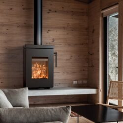 Modern wood-burning stove built into a wooden wall, flames visible. Cozy reading corner with sofa and armchair.
