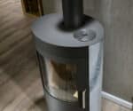 Kachel met spekstenen bekleding en rond kijkvenster, geplaatst in een hoek op een houten vloer. Bovenop een rond metalen deksel.