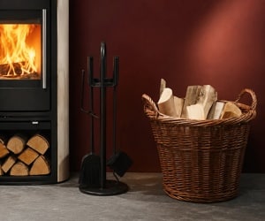 Stove with a burning fire, next to it a basket full of logs and fireplace utensils in front of a red wall.