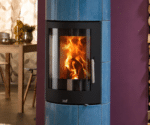 A blue wood-burning stove with a blazing fire behind a large glass window. The stove stands in front of a purple wall, with a table in the background.