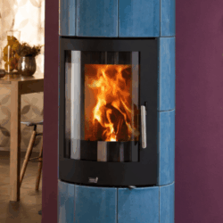 A blue wood-burning stove with a blazing fire behind a large glass window. The stove stands in front of a purple wall, with a table in the background.