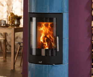 A blue wood-burning stove with a blazing fire behind a large glass window. The stove stands in front of a purple wall, with a table in the background.