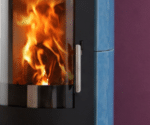 A view of the fire through the panoramic glass panel of a blue wood-burning stove; flames are blazing inside. A stainless steel handle is visible on the side.