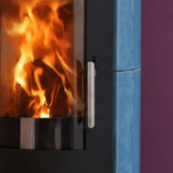 A view of the fire through the panoramic glass panel of a blue wood-burning stove; flames are blazing inside. A stainless steel handle is visible on the side.