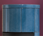The upper part of a blue, cylindrical wood-burning stove in front of a purple wall. Glossy ceramic tiles with vertical grout lines.