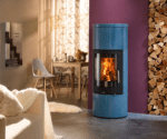 A blue wood-burning stove with a fire burning, in front of a purple wall. A stack of firewood next to it. In the background, a dining area with a table and chairs.
