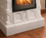 Detailaufnahme eines weißen Kachelofens mit brennendem Feuer hinter einer Glastür. Verzierungen auf den Kacheln, umgeben von Fliesenboden.