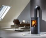 Kaminofen mit Specksteinverkleidung in modernem Wohnzimmer, brennendes Feuer sichtbar. Daneben ein Stuhl, Teppich und Dekoration auf einem Tisch.